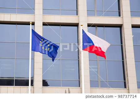 EU flag and Czech Republic flag on flagpole on modern consulate building. Flag of European Union and Czech flag on glass blue facade. EU flag and Czech Republic flag on flagpole on modern consulate building. Flag of European Union and Czech flag on glass blue facade. 98737714