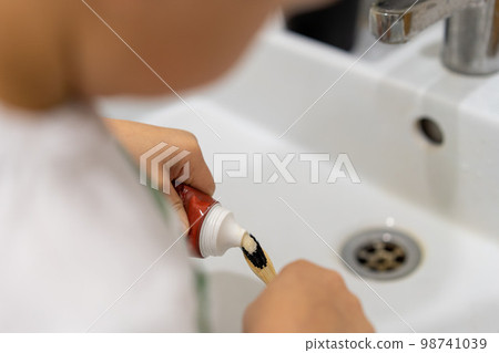 child squeeze tooth paste onto bamboo brush over white sink in bathroom 98741039