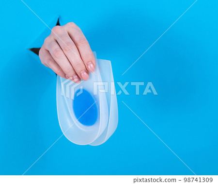 Woman holding orthopedic silicone heel pads. A woman's hand sticks out of a hole in a cardboard blue background. Woman holding orthopedic silicone heel pads. A woman's hand sticks out of a hole in a cardboard blue background. 98741309