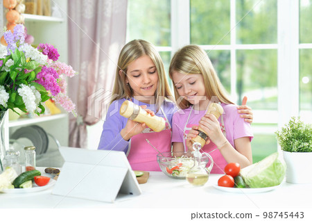 Two girls in pink aprons preparing salad on kitchen with tablet 98745443