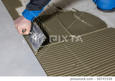 Tiler worker placing or tiling gray ceramic tile in the position over adhesive glue with lash tile leveling system, renovation or recontruction, concept of building 98745598