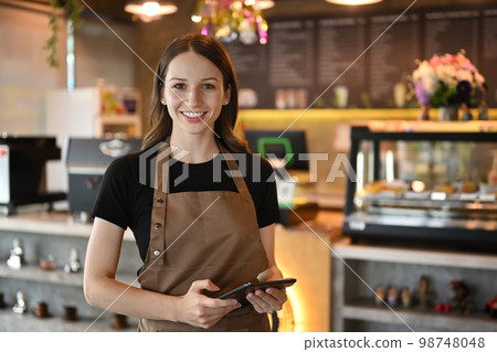 Half length of cheerful caucasian entrepreneur in apron standing in modern cafe interior and using digital tablet Half length of cheerful caucasian entrepreneur in apron standing in modern cafe interior and using digital tablet 98748048