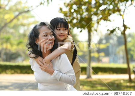 Image of smiling idle age grandmother giving piggyback ride to cute little granddaughter during walking on in the park 98748107