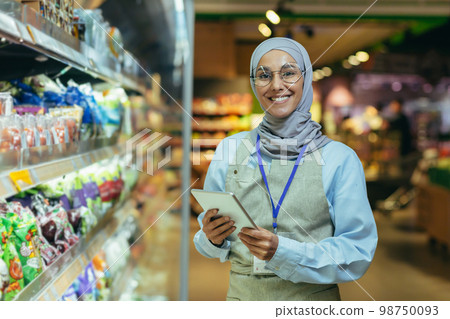 A young Arab woman in a hijab works in a supermarket, salesperson, consultant, manager. Standing with a folder in his hands and a badge in the grocery department. He looks at the camera, smiles. 98750093