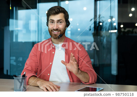 A young man in a red shirt in a headset sits in the office at the table and conducts business training on camera. Explains, advises, smiles. 98750118