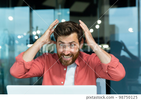 Close-up photo. Shocked and happy man in red shirt sitting at desk in office and looking at laptop. Screams with joy, grabs his head with his hands. 98750125