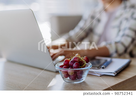 Side view of woman holding bowl with grapes above desk with computer, diary and smartphone. Work-life balance concept. 98752341