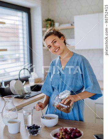 Young woman preparing muesli for breakfast in her kitchen, morning routine and healthy lifestyle concept. 98752415
