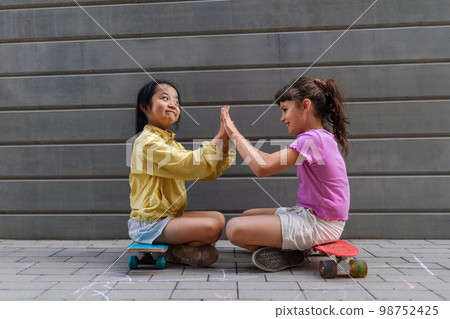 Asian girl with her friend sitting in skateboards in city street, talking and playing, active lifestyle kids concept. Asian girl with her friend sitting in skateboards in city street, talking and playing, active lifestyle kids concept. 98752425