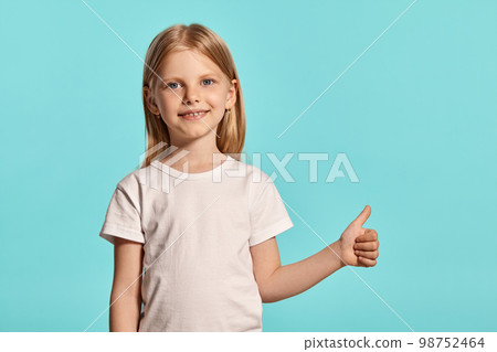 Close-up studio shot of a lovely blonde little girl in a white t-shirt posing against a blue background. 98752464