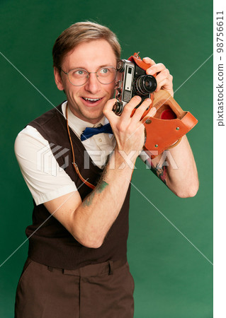 Amateur photographer. Portrait of man in stylish classical clothes, shorts and vest posing over green studio background. Concept of emotions, facial expression, lifestyle Amateur photographer. Portrait of man in stylish classical clothes, shorts and vest posing over green studio background. Concept of emotions, facial expression, lifestyle 98756611
