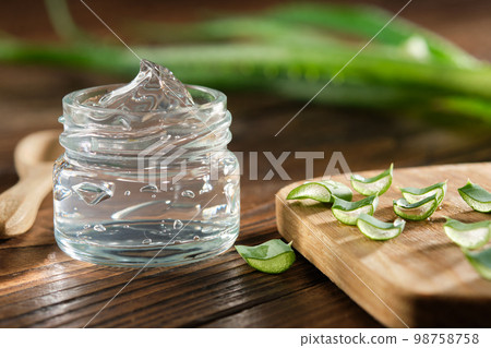Transparent glass jar of aloe vera gel, sliced aloe leaves on a cutting board. Aloe vera plant on background. Natural cosmetic and medicinal remedy. 98758758
