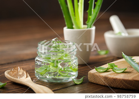 Glass jar of aloe vera gel, sliced aloe plant on a cutting board. Mortars of aloe vera leaves on background. Natural cosmetic and  healing remedy. 98758759