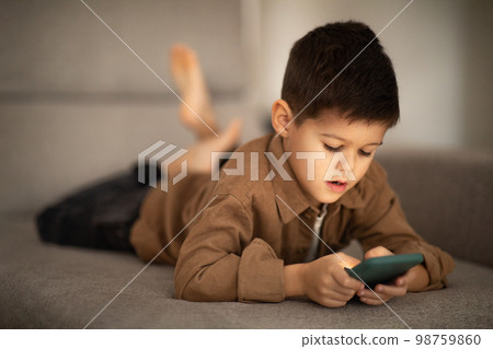 Busy cute small european boy lies on sofa, plays game, watches video on smartphone in room interior, close up Busy cute small european boy lies on sofa, plays game, watches video on smartphone in room interior, close up 98759860