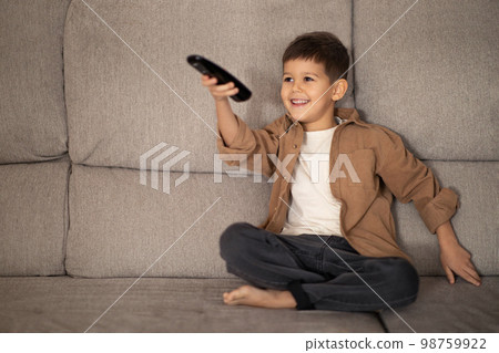 Happy small european boy with remote control watch video, has fun and shoots, switches channels on TV 98759922
