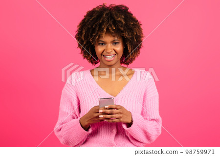 Cheerful young black curly lady in casual with braces typing on smartphone, read message Cheerful young black curly lady in casual with braces typing on smartphone, read message 98759971