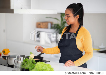 Cheerful millennial african american lady housewife in apron frying fish on plate with organic fresh vegetables 98760040
