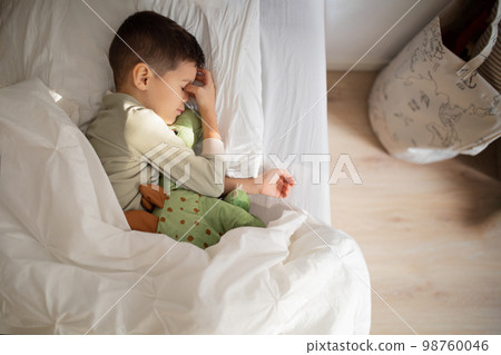 Tired european small kid in pajamas sleeps, wakes up on bed with dinosaur toy in bedroom interior Tired european small kid in pajamas sleeps, wakes up on bed with dinosaur toy in bedroom interior 98760046