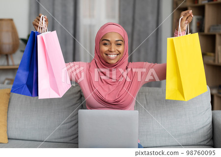 Excited black lady in hijab sitting on sofa with laptop and lifting gift bags above her head, happy to get big discount Excited black lady in hijab sitting on sofa with laptop and lifting gift bags above her head, happy to get big discount 98760095