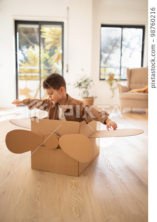 Cheerful small european boy in casual plays in cardboard box helicopter, flies, fantasizes alone in children room Cheerful small european boy in casual plays in cardboard box helicopter, flies, fantasizes alone in children room 98760216