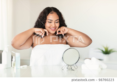 Beautiful body positive lady making lymphatic drainage facial self massage, looking at mirror, sitting in bedroom 98760223