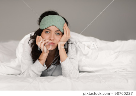 Sad tired young european woman in sleep mask lies on white comfortable bed under covers, wakes up in bedroom Sad tired young european woman in sleep mask lies on white comfortable bed under covers, wakes up in bedroom 98760227