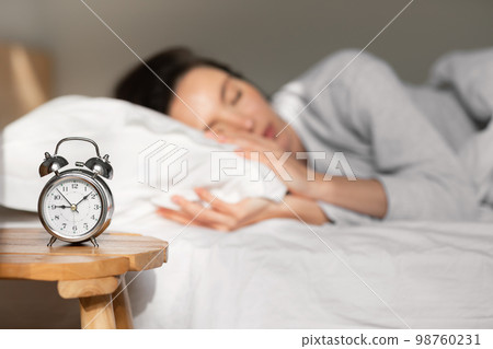 Tired young european lady lies and sleeps on white comfortable bed, selective focus on alarm clock on table Tired young european lady lies and sleeps on white comfortable bed, selective focus on alarm clock on table 98760231
