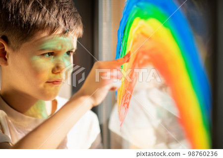 Smiling cute european small kid draws rainbow on window with finger and paint, close up, sun flare 98760236
