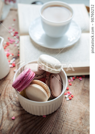 Macaroons in a bowl and a cup of coffee, a book on the background of small hearts on a wooden background, top view 98760252