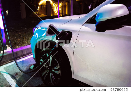 The white car is connected to the electric charging station at night in neon lights in the rain 98760635
