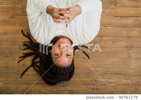 Positive young african american woman chatting on cellphone and smiling on floor at home. Cheerful young woman communicating with someone on smartphone indoors 98761179