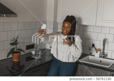 Happy modern African American young girl talking on video chat and sit at home communication concept. Smiling hipster girl making self portrait on her telephone at home 98761190