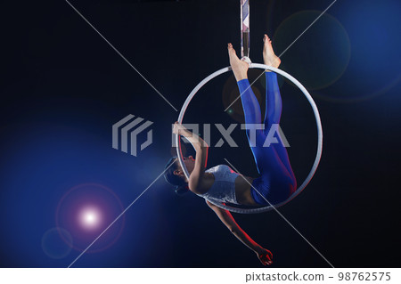Young woman performing acrobatic element on aerial ring against dark background 98762575