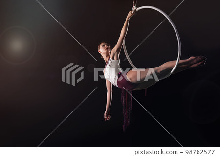 Young woman performing acrobatic element on aerial ring against dark background Young woman performing acrobatic element on aerial ring against dark background 98762577