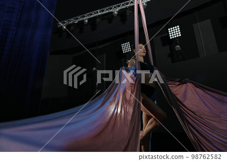 Young woman performing acrobatic element on aerial silk indoors, low angle view 98762582