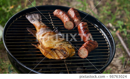 Top view of fresh grilled whole fish, skewer, sausages on round black charcoal grill, green grass background. Barbecue, grill and food concept. Camping with family. Top view of fresh grilled whole fish, skewer, sausages on round black charcoal grill, green grass background. Barbecue, grill and food concept. Camping with family. 98763024