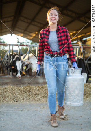 Cheerful girl dairy farm worker carrying can of milk in cowshed Cheerful girl dairy farm worker carrying can of milk in cowshed 98764833