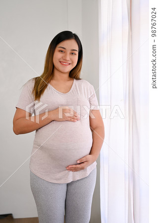 Attractive Asian pregnant woman stands by the window, touching her belly with love and care. Attractive Asian pregnant woman stands by the window, touching her belly with love and care. 98767024