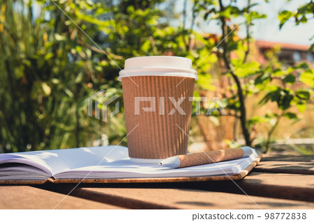 Eco recycling paper cup with coffee or tea on kraft paper with empty paper notebook on wooden bench. Concept of study work outdoors. Take away coffee to go. Copy space for text. Disposable Cardboard 98772838