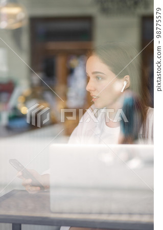 Young woman working with laptop in cafe. View through the window with reflections 98775579