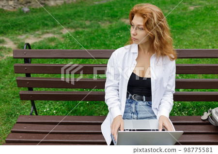 Young beautiful redhead woman using laptop at the bench in a park 98775581