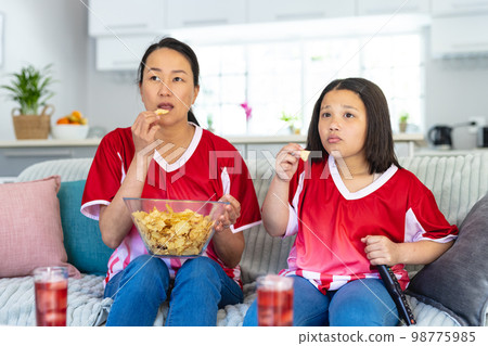 Asian mother and daughter in football shirts watching soccer game, supporting and eating crisps 98775985