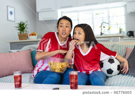 Asian mother and daughter in football shirts watching soccer game, supporting and eating crisps Asian mother and daughter in football shirts watching soccer game, supporting and eating crisps 98776005