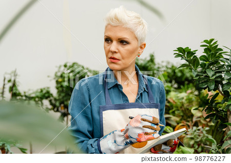 Serious woman gardener checking plants, taking notes about greenery. 98776227