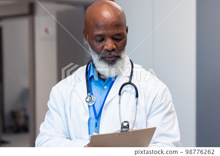 African american senior male doctor reading notes in hospital corridor, copy space African american senior male doctor reading notes in hospital corridor, copy space 98776262