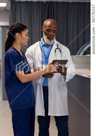 Vertical of diverse female and senior male doctor using tablet and talking in hospital, copy space 98776337