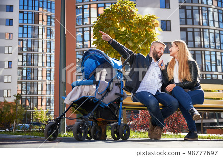 Young parents sitting on bench with unrecognizable baby in stroller, having rest in city. Front view of father raising hand, pointing at new apartment in new city district. Concept of family. 98777697