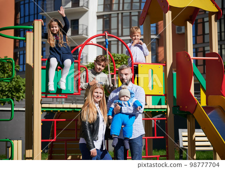 Happy family - father, mother and children having fun together on playground. Three cheerful kids playing while parents holding toddler in arms. Modern residential buildings on background. 98777704