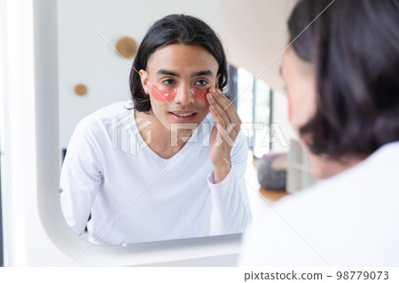 Happy biracial man looking in mirror, applying eye patches in bathroom 98779073