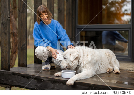 Young woman feeds her cute white dog on porch of a house 98780678